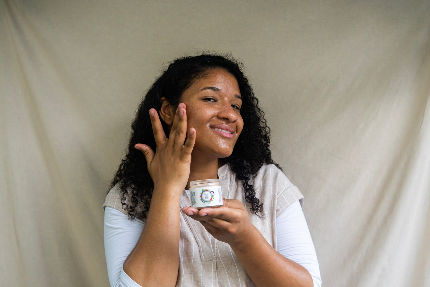 A smiling woman holds a jar of the Sweet Dreams Glow Balm while applying it to her face.